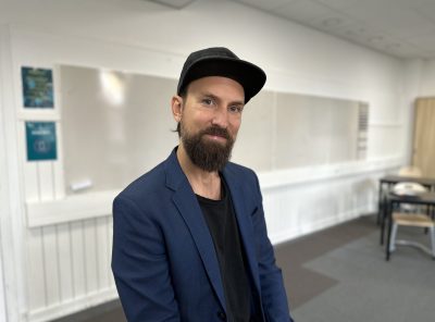 Läraren Erik sitter i ett klassrum framför en whiteboard-tavla. Han har på sig en mörkblå kavaj och svart keps.