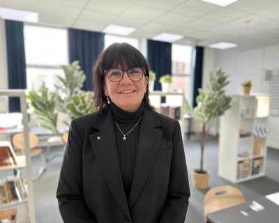 Rektor Pernilla står i skolans nya bibliotek. Hon har på sig en svart kavaj och glasögon.