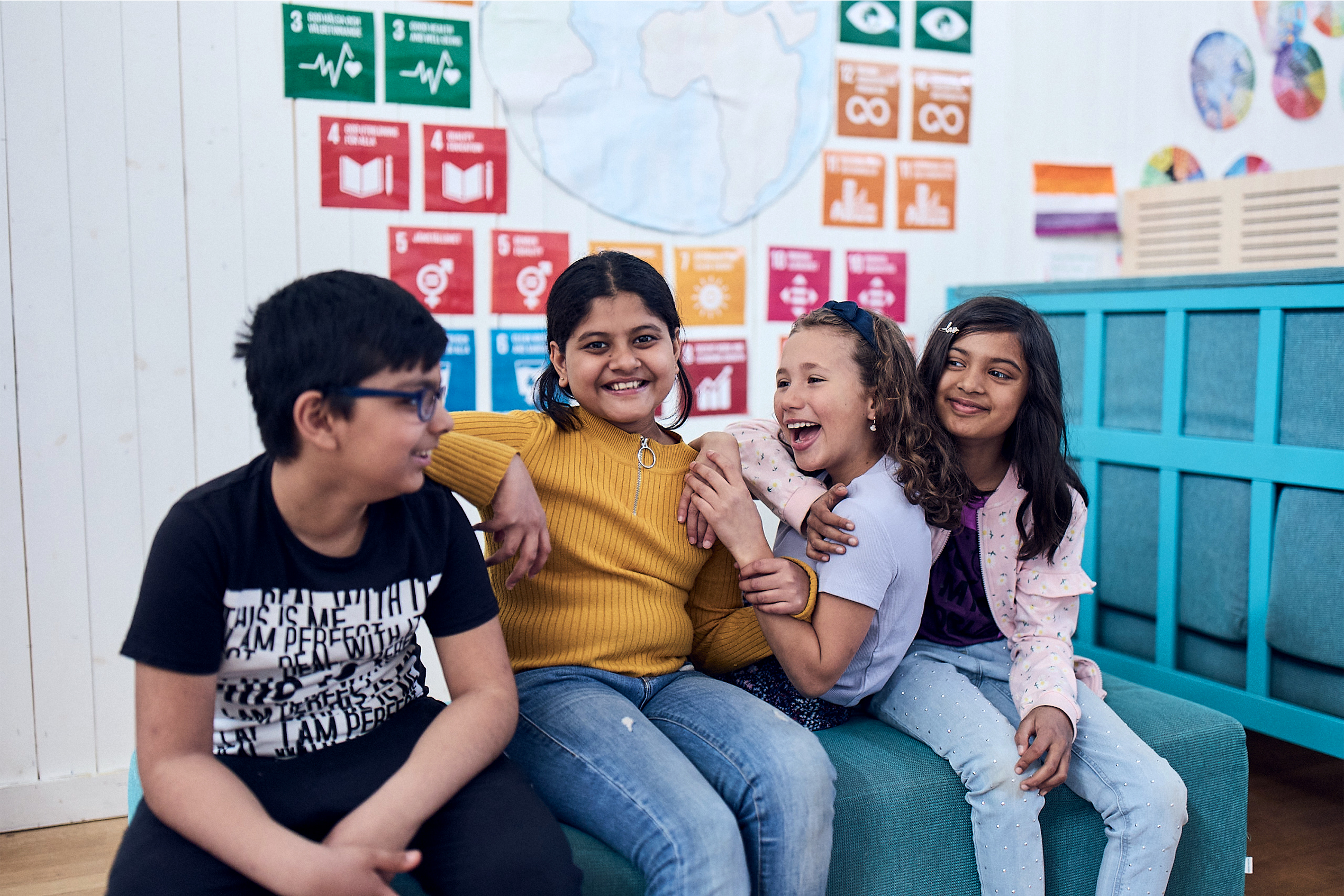 Fyra glada elever sitter på en bänk på Vittra med FN:s Globala mål i bakgrunden.