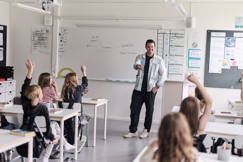 Lärare står i klassrum vid tavla med elever som räcker upp handen