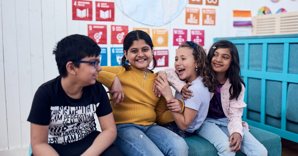 Fyra glada elever sitter på en bänk på Vittra, i bakgrunden syns FN:s globala mål.