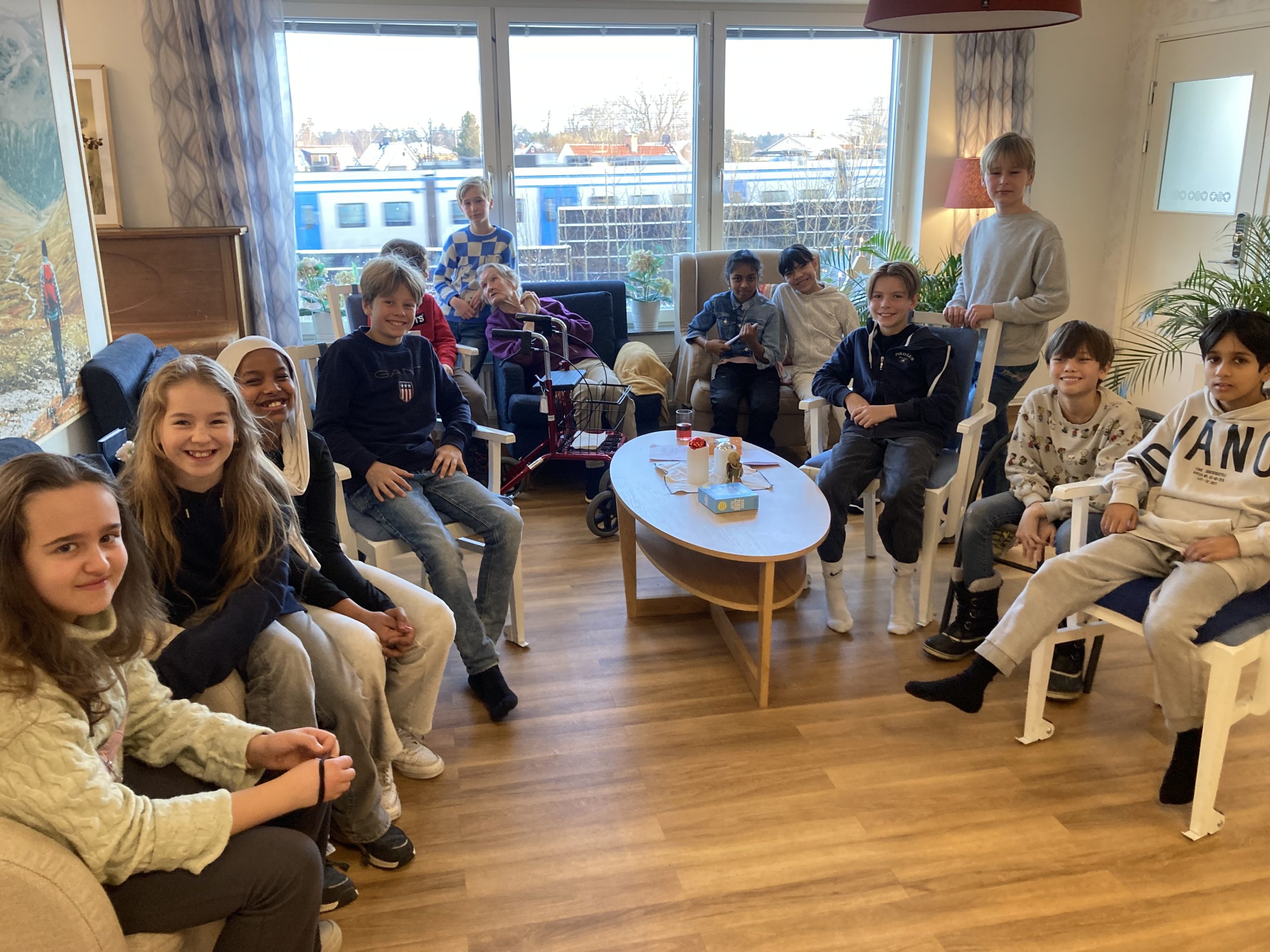 Elever från Vittra Rösjötorp sitter på stolar i en halvmåne. Mitt i sitter en kvinna från äldreboendet Attento.