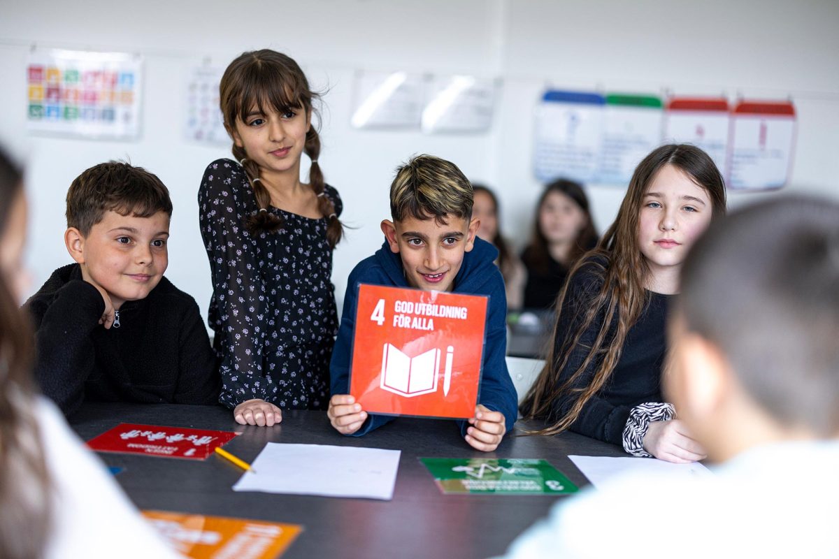 Elever sitter vid skolbänken och en av dem håller i en skylt där det står "God utbildning för alla"