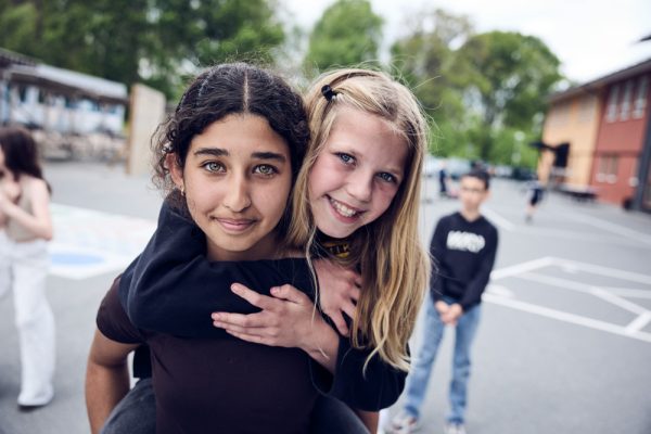 En elev har en annan på ryggen. De ler och ser in i kameran.